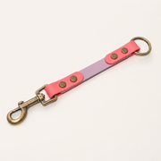 A double-colored BioThane® strap accessory with a bright pink outer layer, a lavender purple inner layer, brass rivets, and antique bronze hardware including a lobster clasp and a ring, shown on a plain off-white background.