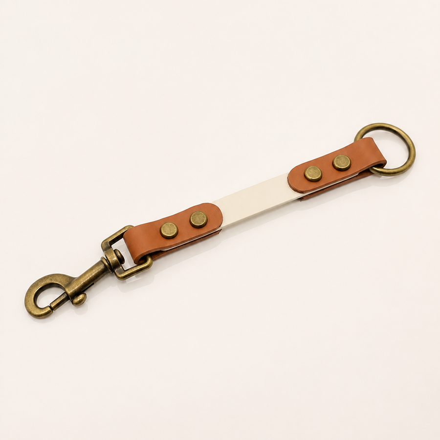 A BioThane® strap accessory with brass-toned hardware, featuring a swivel snap hook on one end, a ring on the other, and two brass rivets on a tan BioThane® section over a white strap, photographed on a light beige background.